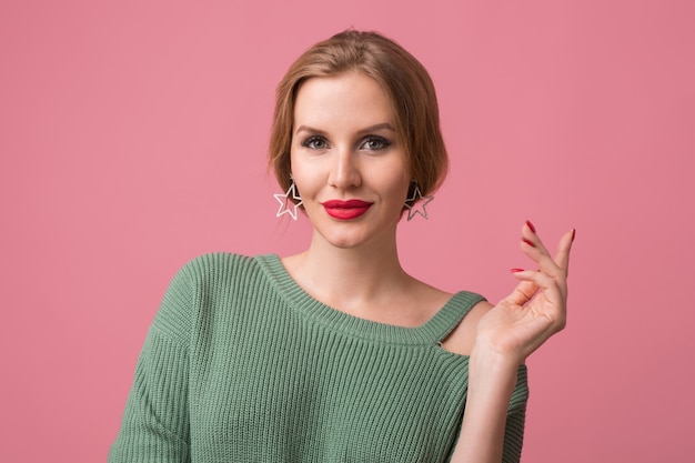 Close-up portret młodej seksownej atrakcyjnej kobiety, stylowy makijaż, czerwone usta, zielony sweter, modelka pozowanie w studio, na białym tle, różowe tło, kolczyki, patrząc w kamerę, trzymając rękę w górze, elegancki