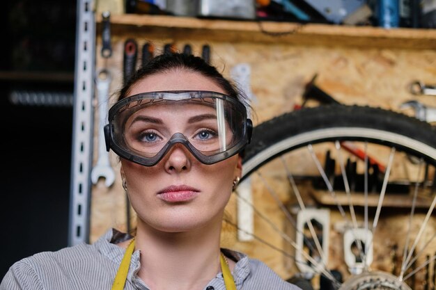 Close-up portret pięknej brunetki kobiet noszących ubrania robocze, fartuch i gogle, stojąc w warsztacie przed narzędziami ściennymi.