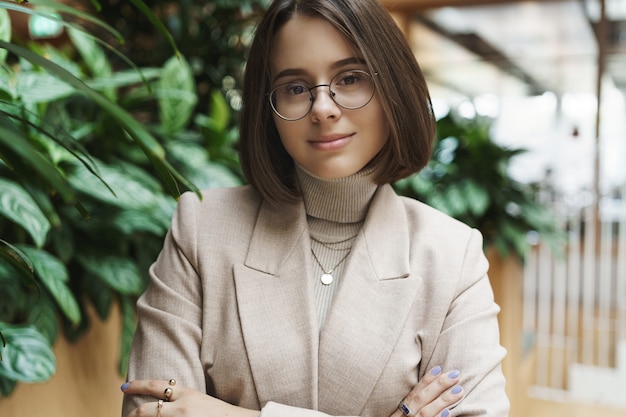 Close-up portret stylowej, ładnej młodej kobiety, wyglądaj profesjonalnie, skrzyżuj ręce w klatce piersiowej i uśmiechnięty pewny aparat, noś okulary, stojąc w pobliżu recepcji lub holu biurowego, omawiaj biznes.