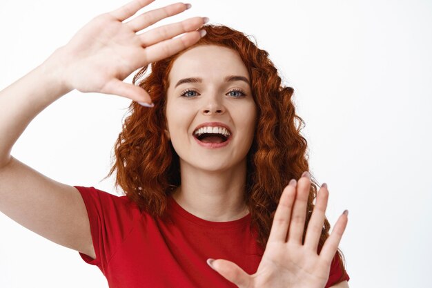 Close-up portret szczęśliwej rudej kobiety wykonującej gest ramek rąk, spójrz na zdjęcie lub obrazowanie czegoś, stojąc w koszulce na białej ścianie
