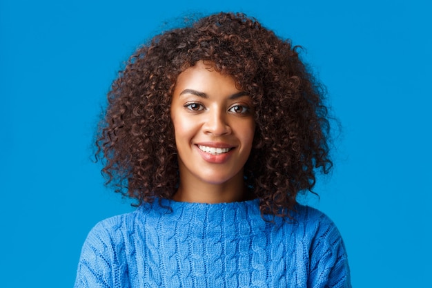 Close-up portret urocza młoda afroamerykańska kobieta z kręconymi fryzurami w stylu afro, uśmiechnięta z radosnym przyjemnym wyrazem, ciesząca się zimowymi wakacjami, ubrana w sweter, niebieska ściana.