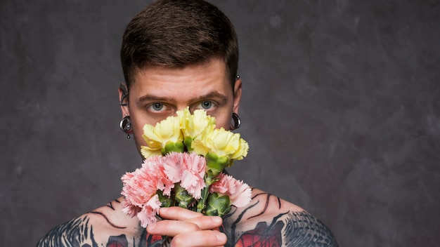 Close-up tatuaż młody człowiek z przebitymi uszami gospodarstwa różowe i żółte kwiaty goździka przed jego ustami