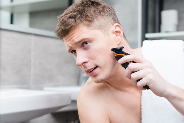 Bezpłatne zdjęcie close-up uśmiechniętego młodego człowieka golenie z elektryczną maszynką do golenia w łazience