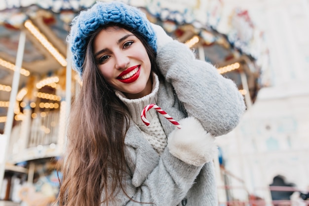 Close-up zdjęcie uroczej dziewczyny z czarnymi włosami i czerwonymi ustami chłodzi zewnątrz z bożonarodzeniowym lizakiem. Portret śmiejącej się młodej kobiety w niebieskim berecie z dzianiny pozowanie w parku rozrywki w grudniu.