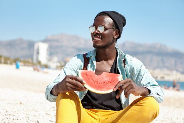 Co za wspaniały dzień! Szczęśliwy młody turysta Afro amerykański na sobie modne ubrania jedzenie dojrzałego arbuza, siedząc na plaży z niewyraźne miasto