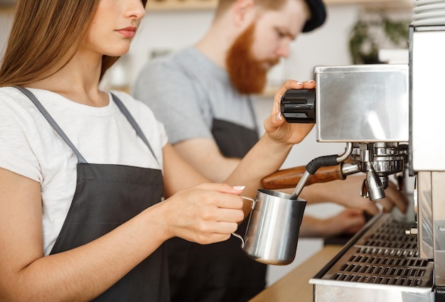 Coffee Business Concept portret lady baristy w fartuchu przygotowującym i gotującym na parze mleko na kawę ze swoim partnerem, stojąc w kawiarni