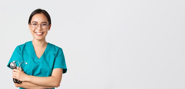Covid-19, choroba koronawirusowa, koncepcja pracowników opieki zdrowotnej. Pewna szczęśliwa azjatycka lekarka, lekarz w okularach, skrzyżowane ramiona w klatce piersiowej i uśmiechnięta, trzymająca stetoskop do badania