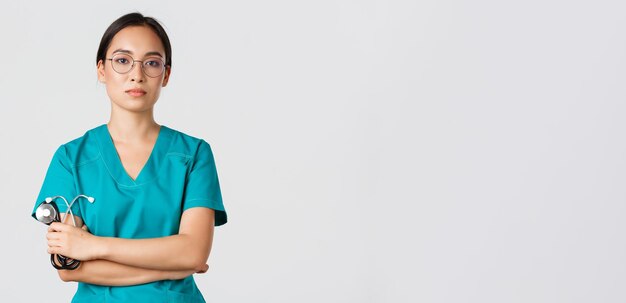 Covid-19, choroba koronawirusowa, koncepcja pracowników opieki zdrowotnej. Profesjonalna pewna siebie, poważnie wyglądająca lekarka, lekarz w okularach i zaroślach, trzymająca stetoskop, stojąca na białym tle.