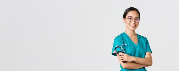 Covid-19, choroba koronawirusowa, koncepcja pracowników opieki zdrowotnej. Profesjonalny przystojny azjatycki lekarz, pracownik medyczny w okularach i zaroślach, skrzyżowane ramiona i uśmiechnięte, białe tło.