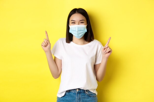 Covid-19, koncepcja pandemii i dystansu społecznego. młoda azjatycka kobieta w białej koszulce i masce medycznej z koronawirusa, wskazując palcami w górę, pokazując specjalną ofertę