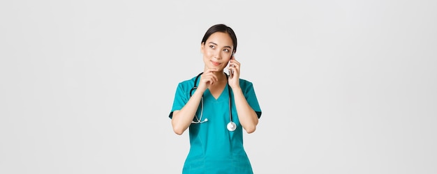 Covid-19, pracownicy służby zdrowia i koncepcja zapobiegania wirusom. portret uśmiechniętej azjatyckiej lekarki, stażystka w zaroślach rozmawiająca przez telefon i wyglądająca na zamyśloną, myślącą lub dokonującą wyboru