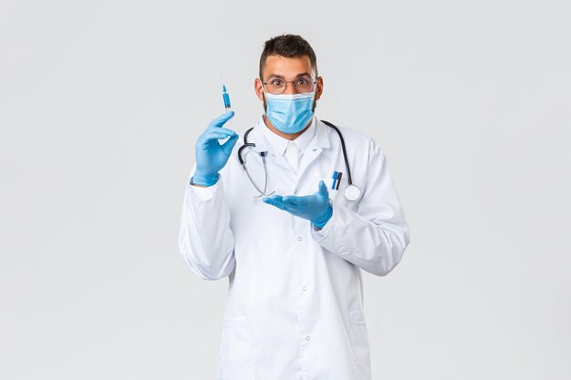 Covid-19, pracownicy służby zdrowia, koncepcja pandemii i zapobiegania wirusom. Podekscytowany lekarz w białym fartuchu, masce medycznej i rękawiczkach, wprowadza nową szczepionkę, zdumiony wskazując na strzykawkę