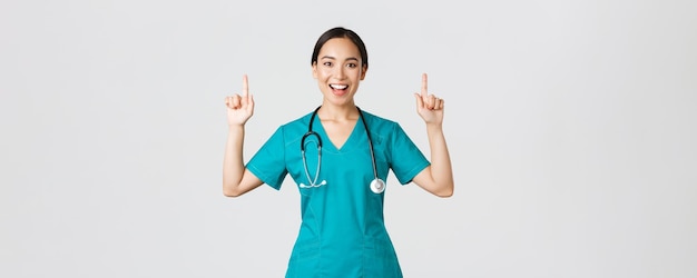 Covid-19, pracownicy służby zdrowia, koncepcja pandemii. uśmiechnięta, wesoła ładna pielęgniarka, lekarka lub stażystka w fartuchu, wskazując palcami w górę, pokazując baner, ogłaszając ogłoszenie, demonstrując reklamę