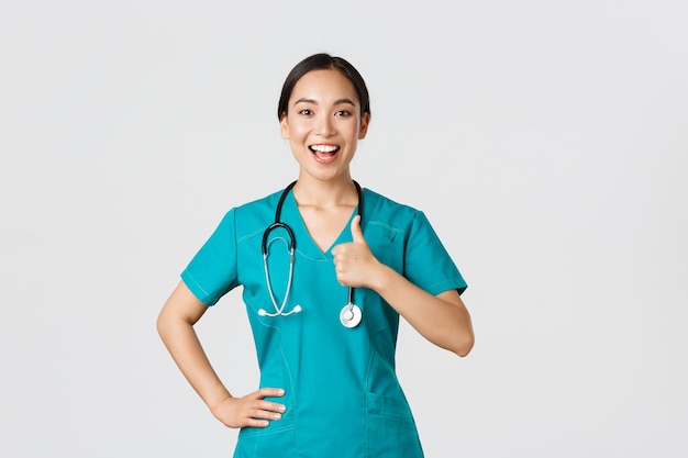 Covid19 pracownicy opieki zdrowotnej koncepcja pandemiczna Uśmiechnięta optymistyczna, pewna siebie pielęgniarka azjatycka w zaroślach ma wszystko pod kontrolą, pokazuje kciuk w górę w zatwierdzeniu, gwarantuje jakość usług w klinice