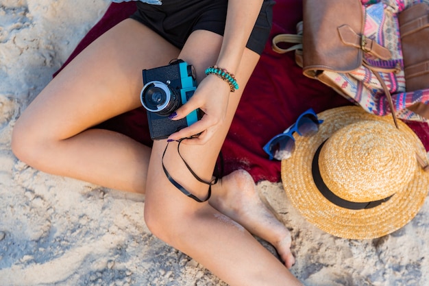 Cudowna kobieta o opalonym ciele, pełnych czerwonych ustach i długich nogach pozuje na tropikalnej słonecznej plaży. Nosi krótki top, szorty i słomkowy kapelusz.