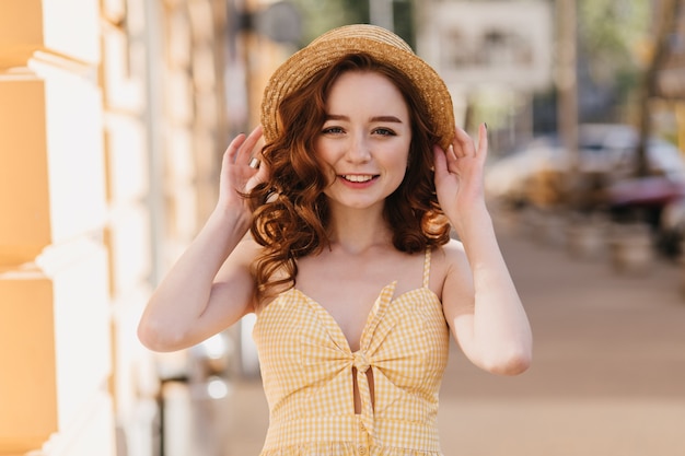 Cudowna ruda kobieta w żółtym stroju vintage idąca ulicą. Odkryte zdjęcie rozmarzonej białej dziewczyny w słomkowym kapeluszu.