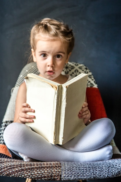 Bezpłatne zdjęcie cute dziewczynka siedzi na pięknym krześle z książką w ręku, pojęcie edukacji i życia szkolnego