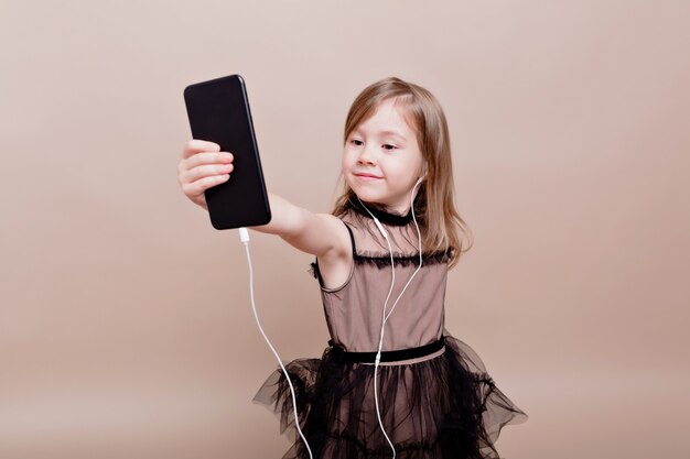 Cute little girl zabawy i bierze selfie. Mała dziewczynka zaskoczona, patrząc w telefon i uśmiecha się na odizolowanej ścianie, prawdziwe emocje, dobry nastrój, stylowa mała dziewczynka
