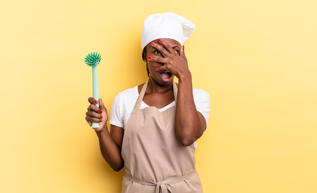 Czarna kobieta kucharz afro wygląda na zszokowaną, przestraszoną lub przerażoną, zakrywając twarz dłonią i zerkając między palcami. koncepcja czyszczenia naczyń
