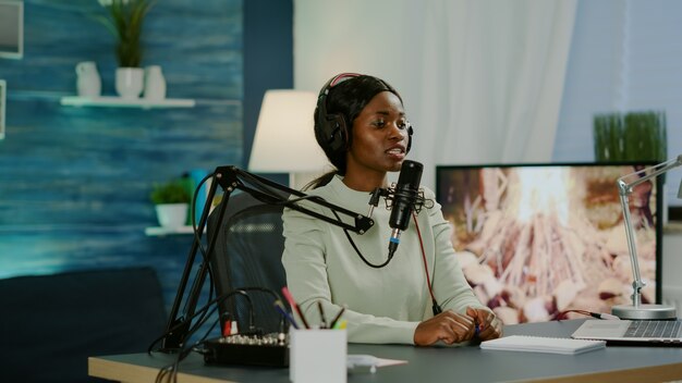 Czarna kobieta prowadząca show online patrząca w laptopie rozmawiająca w mikrofonie podcastowym z rozrywką słuchaczy. Przemawiając podczas transmisji na żywo, bloger dyskutujący na vlogu w słuchawkach.