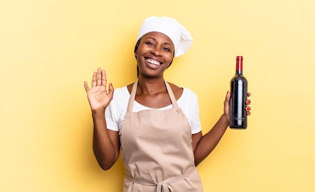 Czarna kobieta szefa kuchni afro uśmiechnięta radośnie i radośnie, machająca ręką, witająca i witająca cię lub żegnająca. koncepcja butelki wina