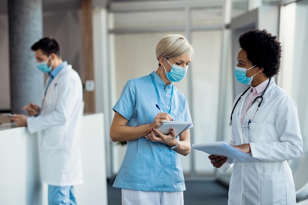 Czarna lekarka i pielęgniarka z maskami na twarzy komunikują się na korytarzu szpitala