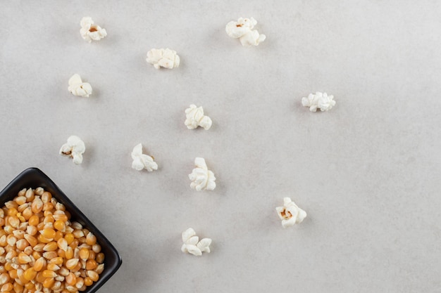 Czarna miska wypełniona ziarnami kukurydzy obok porozrzucanych kawałków popcornu na marmurowym stole.
