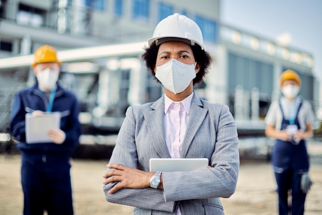Czarna pewna siebie kobieta stojąca ze skrzyżowanymi rękami podczas noszenia maski na placu budowy
