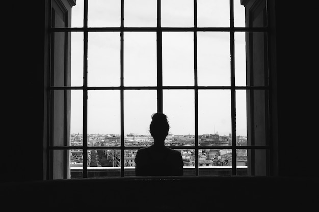 Czarno-białe ujęcie samotnej kobiety stojącej przed oknami spoglądającymi na budynki