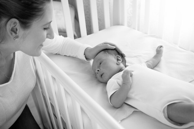 Czarno-biały portret szczęśliwej uśmiechniętej matki pieszczącej swoje dziecko leżące w łóżeczku