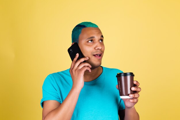 Czarny afrykański mężczyzna w dorywczo na żółtej ścianie niebieskie włosy, ciesząc się filiżanką kawy, pozytywne szczęśliwe emocje rozmawia przez telefon, uśmiech i śmiech