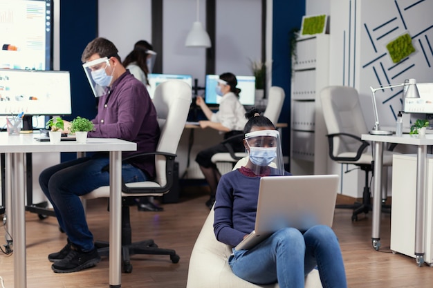 Czarny freelancer z maską ochronną przed koronawirusem siedzący na fotelu pośrodku gabinetu analizujący projekt na cyfrowym tablecie. Wieloetniczny zespół biznesowy pracujący z poszanowaniem dystansu społecznego