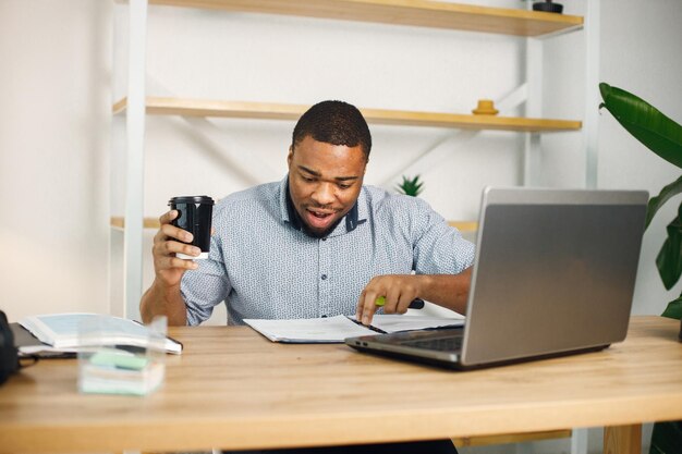 Czarny mężczyzna przedsiębiorca siedzi w biurze przy laptopie i pije kawę
