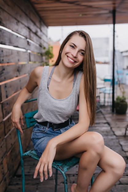 Czarująca blada kobieta siedzi na krześle na miejskiej ulicy i uśmiecha się. Niesamowita brązowowłosa dziewczyna w dżinsowej spódnicy spędza wiosenny poranek w ulicznej kawiarni.