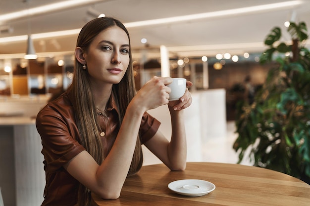 Czas cieszyć się świeżo zaparzoną kawą Atrakcyjna zrelaksowana młoda kobieca kobieta w brązowej sukience siedząca samotnie w kawiarni trzymająca filiżankę i popijając cappuccino w spokojnym przytulnym miejscu uśmiechnięta kamera