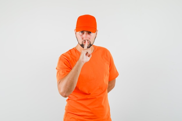 Człowiek dostawy pokazując gest ciszy w pomarańczowy t-shirt, czapkę i patrząc ostrożnie. przedni widok.