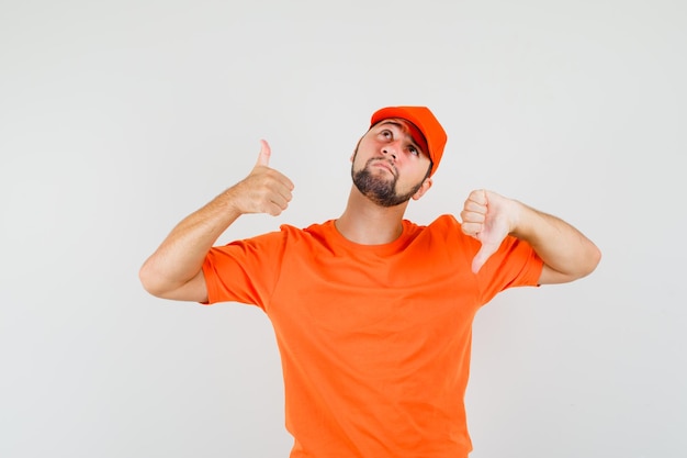 Bezpłatne zdjęcie człowiek dostawy pokazując kciuk w górę iw dół w pomarańczowy t-shirt, czapkę i patrząc zdezorientowany. przedni widok.