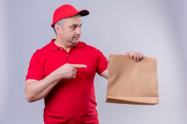 Człowiek dostawy w czerwonym mundurze i czapce, trzymając pakiet papieru, wskazując palcem wskazującym na to, patrząc na bok z poważną twarzą stojącą nad odizolowaną białą przestrzenią
