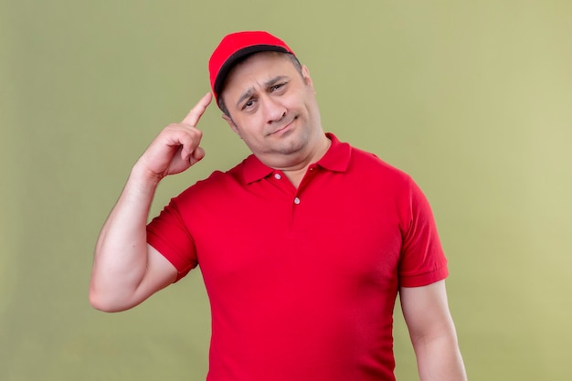Bezpłatne zdjęcie człowiek dostawy w czerwonym mundurze i czapce wskazuje świątynię z palcem mocno koncentrując się na pomyśle stojącym nad zieloną przestrzenią