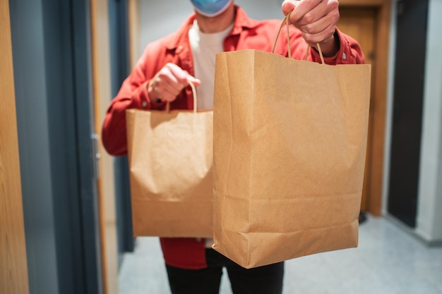 Człowiek dostawy w masce ochronnej trzymając papierową torbę z jedzeniem w wejściu. kurier wręcza klientowi pudełko ze świeżymi warzywami i owocami