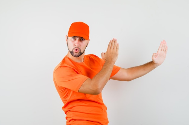 Człowiek dostawy w pomarańczowy t-shirt, czapka pokazująca gest karate chop i patrząc pewnie, widok z przodu.