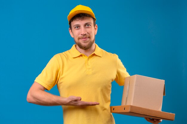 człowiek dostawy w żółtej koszulce polo i czapce, trzymając kartony uśmiechnięty przyjaźnie i prezentujący z dłonią, stojąc na niebiesko