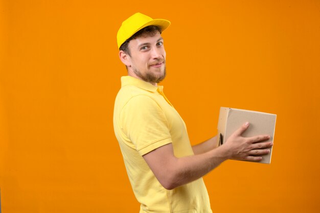 człowiek dostawy w żółtej koszulce polo i czapce, trzymając pakiet box uśmiechnięty przyjazny stojący bokiem na pomarańczowo