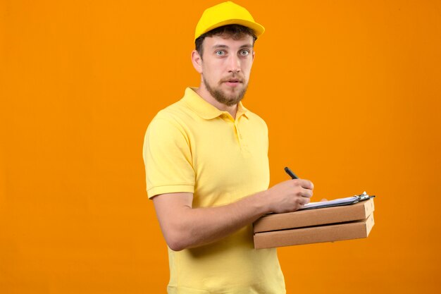 człowiek dostawy w żółtej koszulce polo i czapce, trzymając pudełka po pizzy i schowek, pisząc coś patrząc na kamery z poważną twarzą stojącą na odizolowanych pomarańczy