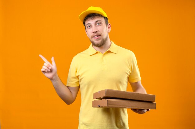 człowiek dostawy w żółtej koszulce polo i czapce, trzymając pudełka po pizzy wskazując palcem w bok uśmiechnięty przyjazny stojący na pomarańczowo