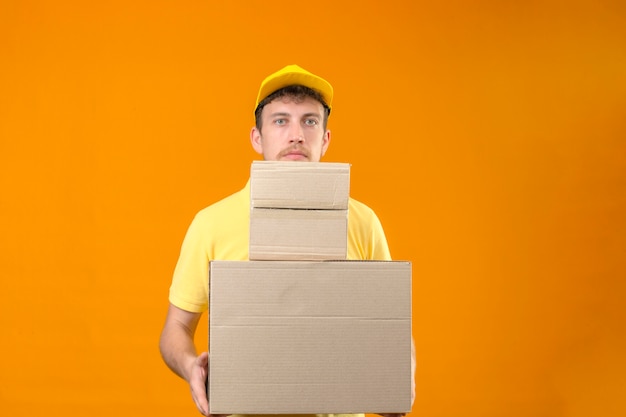 człowiek dostawy w żółtej koszulce polo i czapce trzymającej duże ciężkie pudła wyglądające na zmęczonego i znudzonego stojącego na odizolowanej pomarańczy
