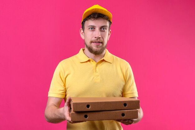 człowiek dostawy w żółtej koszulce polo i czapce trzymającej pudełka po pizzy wyglądający pewnie stojąc na odizolowanym różu