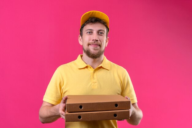 człowiek dostawy w żółtej koszulce polo i czapce trzymający pudełka po pizzy wyglądający pewnie i dumnie uśmiechnięty wesoły stojący na odizolowanym różu