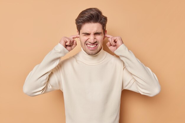 człowiek zatyka uszy nie chce słyszeć denerwującego dźwięku unika hałasu frons twarz nosi casualowy sweter na beżowym nie chce czegoś słuchać. Wyłącz głośność.