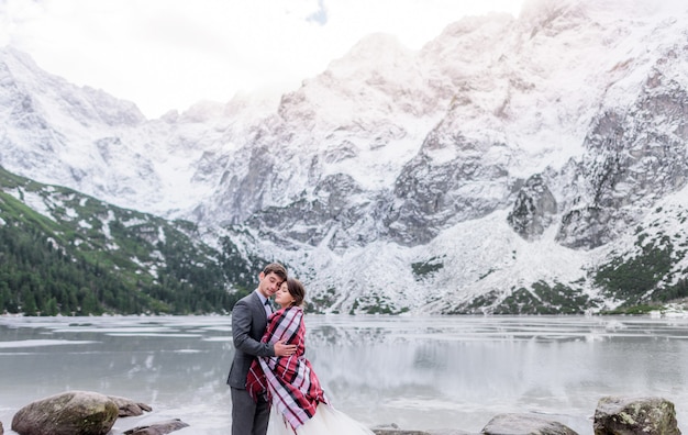 Czuła para przykryta zakochanym kocem marznie w zimowych górach przed zamarzniętym jeziorem